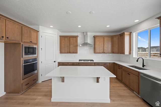kitchen featuring sink, stainless steel appliances, wall chimney range hood, a kitchen island, and light wood-type flooring