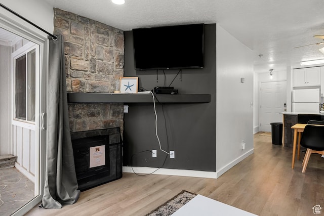 living room with light wood-style floors, a stone fireplace, a textured ceiling, baseboards, and a ceiling fan