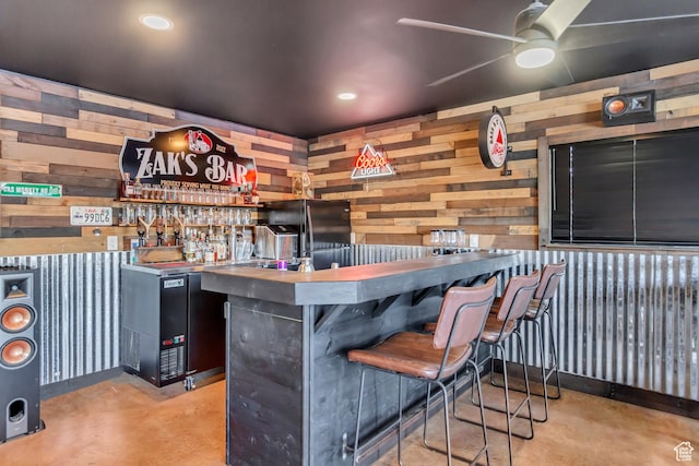 indoor dry bar with concrete floors, wood walls, a ceiling fan, and freestanding refrigerator