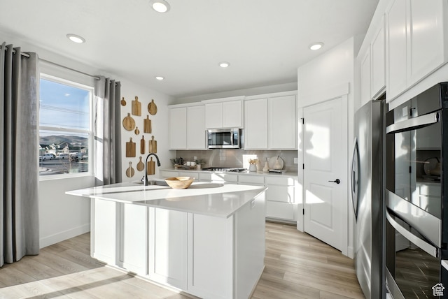 kitchen with light countertops, appliances with stainless steel finishes, an island with sink, white cabinetry, and backsplash