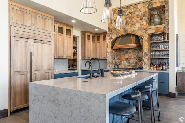 kitchen featuring an island with sink, light stone countertops, custom exhaust hood, a sink, and dark wood-style floors