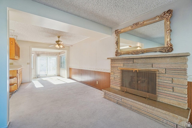 unfurnished living room featuring a stone fireplace, wooden walls, ceiling fan, a textured ceiling, and light colored carpet