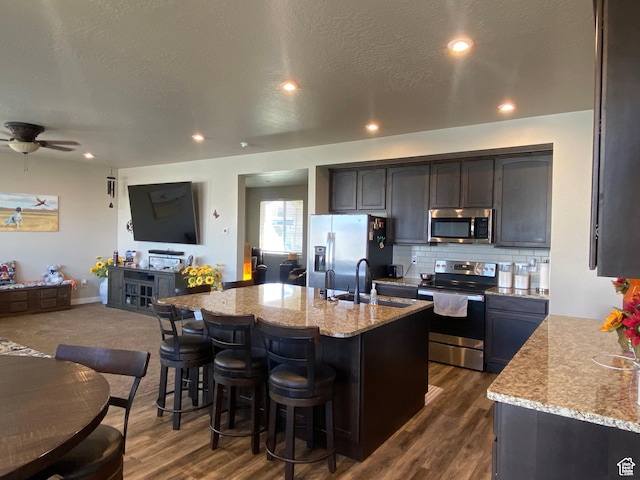 kitchen with dark hardwood / wood-style flooring, light stone counters, a kitchen island with sink, and appliances with stainless steel finishes
