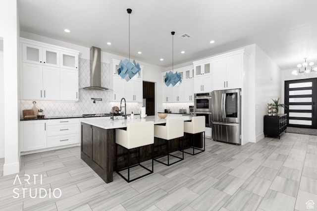 kitchen featuring stainless steel appliances, wall chimney range hood, decorative light fixtures, a kitchen bar, and a kitchen island with sink