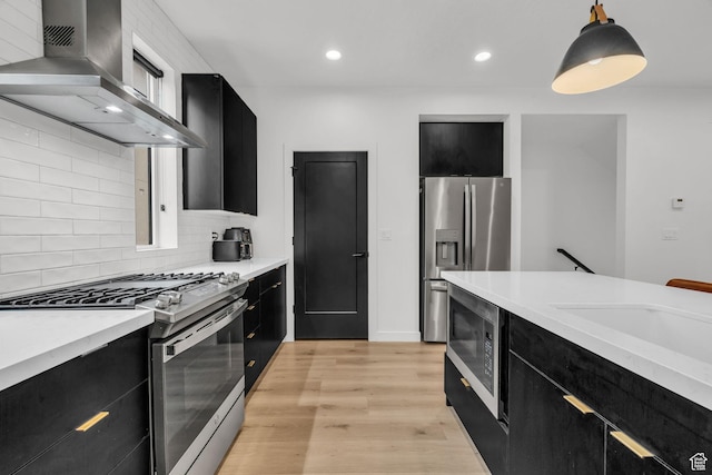 kitchen with wall chimney range hood, decorative light fixtures, light countertops, appliances with stainless steel finishes, and dark cabinets