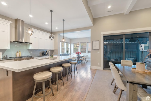 kitchen with a sink, wall chimney range hood, light wood finished floors, a center island with sink, and decorative backsplash