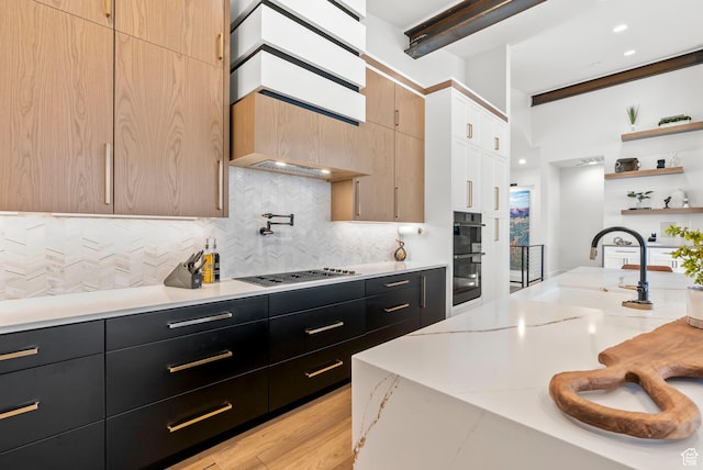 kitchen with light stone countertops, light brown cabinetry, decorative backsplash, black double oven, and light hardwood / wood-style floors