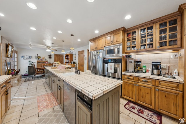 kitchen featuring a kitchen island with sink, hanging light fixtures, stainless steel appliances, tile counters, and light tile patterned flooring