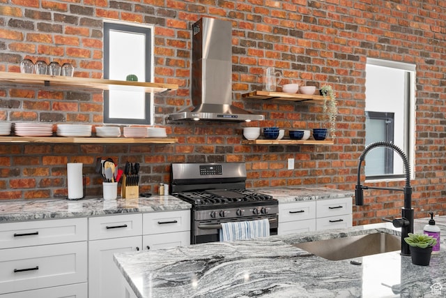 kitchen featuring light stone countertops, brick wall, stainless steel range with gas stovetop, and wall chimney range hood