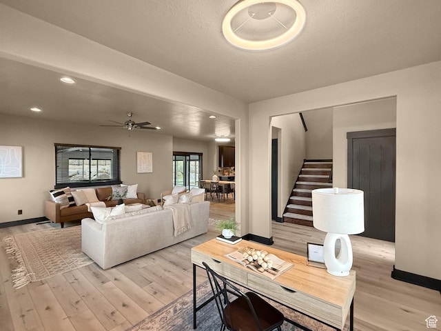 living room featuring a ceiling fan, light wood-style flooring, baseboards, and stairs