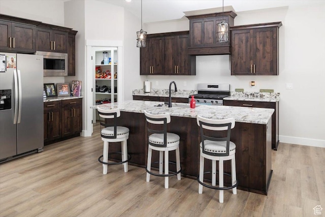 kitchen featuring dark brown cabinetry, decorative light fixtures, stainless steel appliances, light stone counters, and a center island with sink