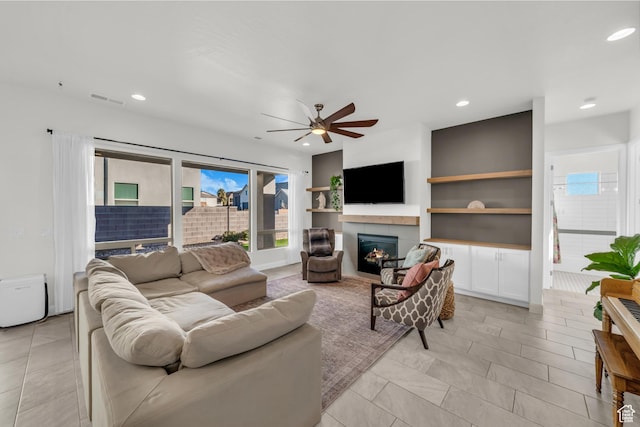 living room with ceiling fan, a tiled fireplace, and built in shelves