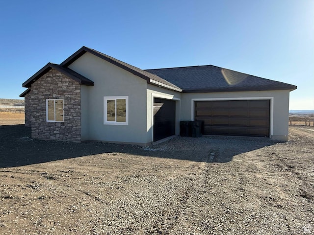 view of front of home with stone siding, dirt driveway, a garage, roof with shingles, and stucco siding
