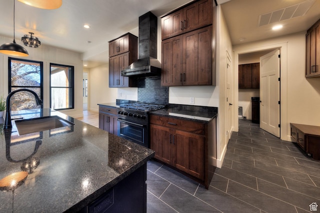 kitchen with pendant lighting, wall chimney range hood, dark stone counters, sink, and gas range oven