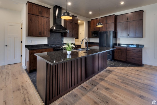 kitchen with sink, black fridge, pendant lighting, an island with sink, and wall chimney exhaust hood