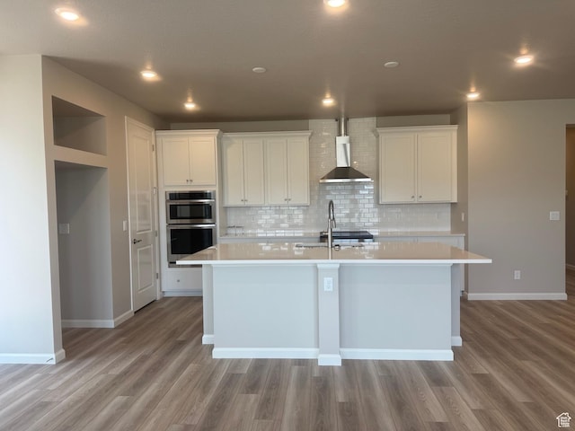 6158 W 11800 S Unit 504, South Jordan, UT 84009 - kitchen featuring wall chimney exhaust hood, double oven, an island with sink, light countertops, and decorative backsplash
