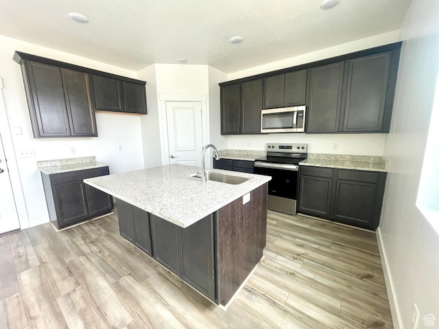 kitchen with light stone counters, appliances with stainless steel finishes, a sink, light wood-style floors, and an island with sink