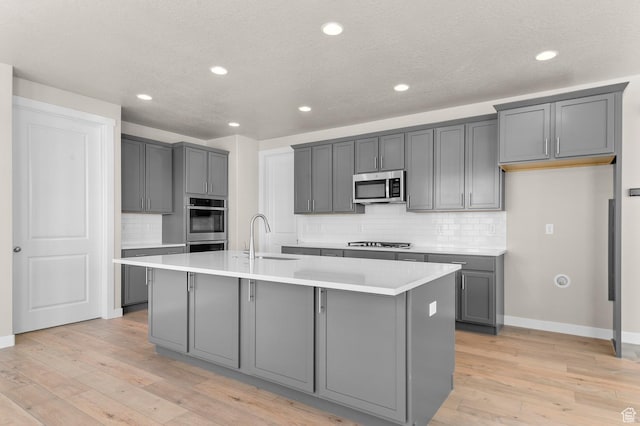 kitchen with gray cabinetry, an island with sink, light wood-style floors, and appliances with stainless steel finishes