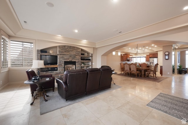 tiled living room with plenty of natural light, a stone fireplace, a raised ceiling, and a notable chandelier