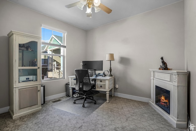office featuring baseboards, a glass covered fireplace, a ceiling fan, and light colored carpet