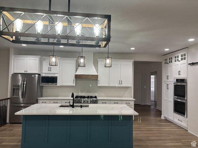 kitchen with dark wood-style flooring, custom exhaust hood, appliances with stainless steel finishes, white cabinets, and an island with sink