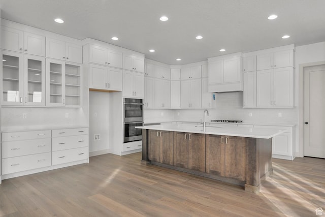 kitchen featuring an island with sink, stainless steel double oven, white cabinets, and light wood-type flooring