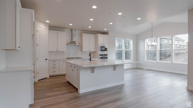 kitchen with stainless steel microwave, an island with sink, light wood-style floors, wall chimney range hood, and an inviting chandelier