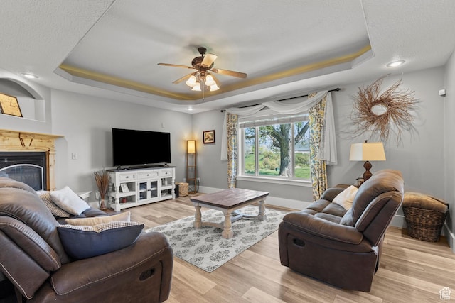 living area featuring light wood finished floors, a ceiling fan, a tray ceiling, baseboards, and a glass covered fireplace