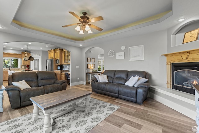living area with a ceiling fan, light wood finished floors, a glass covered fireplace, arched walkways, and a raised ceiling