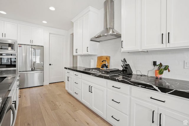 kitchen with appliances with stainless steel finishes, white cabinets, dark stone counters, light hardwood / wood-style floors, and wall chimney exhaust hood