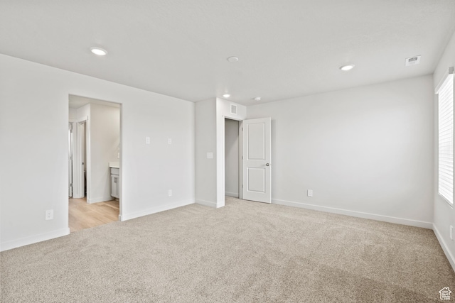 unfurnished bedroom featuring light colored carpet, recessed lighting, baseboards, ensuite bath, and multiple windows