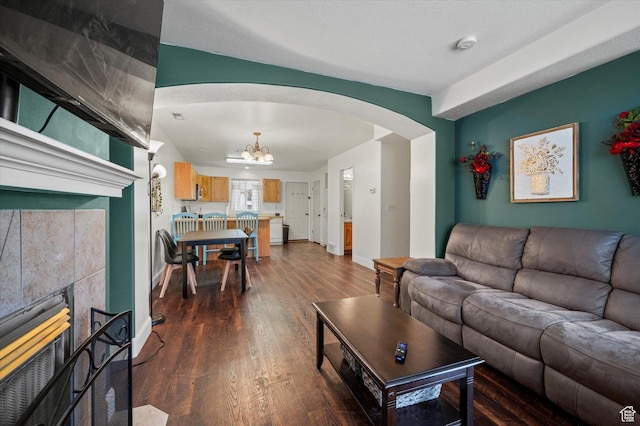 living area featuring arched walkways, a tile fireplace, dark wood-type flooring, baseboards, and an inviting chandelier