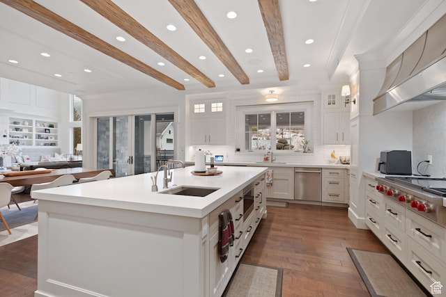 kitchen featuring sink, appliances with stainless steel finishes, beam ceiling, white cabinets, and a center island with sink