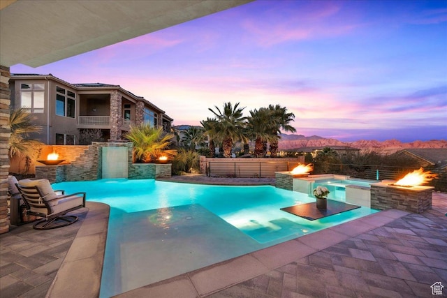 pool at dusk with a jacuzzi, pool water feature, an outdoor fire pit, a mountain view, and a patio area
