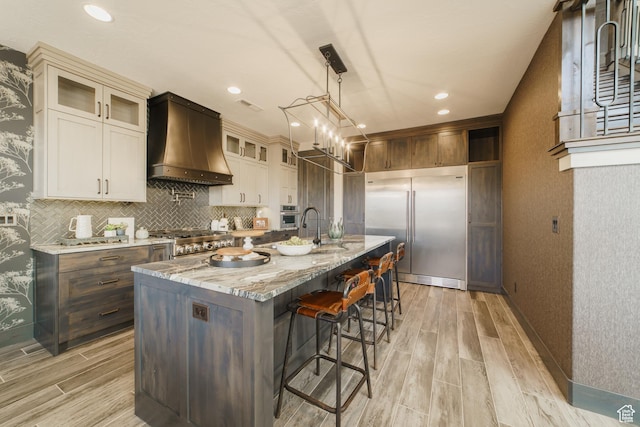 kitchen featuring premium range hood, stainless steel built in refrigerator, light stone counters, a kitchen island with sink, and stove