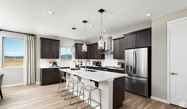 kitchen featuring decorative light fixtures, dark brown cabinets, appliances with stainless steel finishes, a kitchen island with sink, and wall chimney range hood
