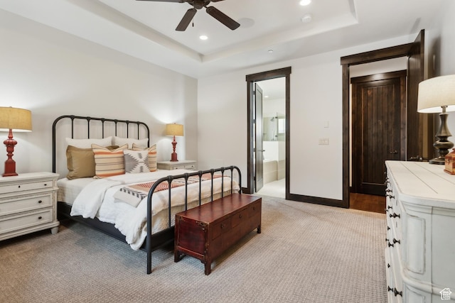 bedroom with a raised ceiling, ensuite bathroom, light colored carpet, and ceiling fan