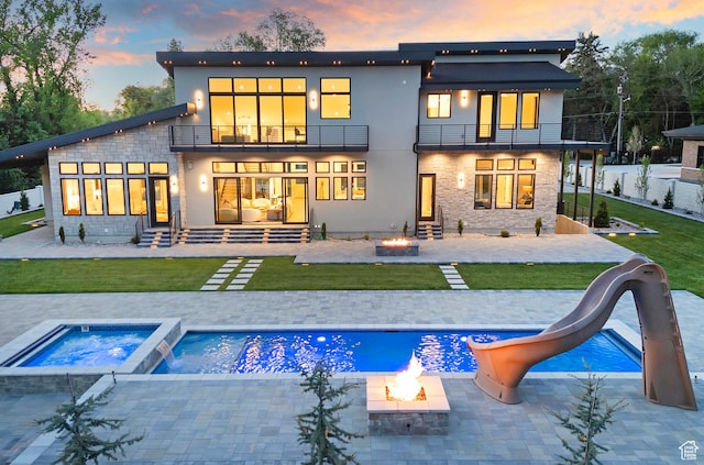 back of property at dusk featuring a fenced in pool, an outdoor fire pit, a lawn, a balcony, and a patio