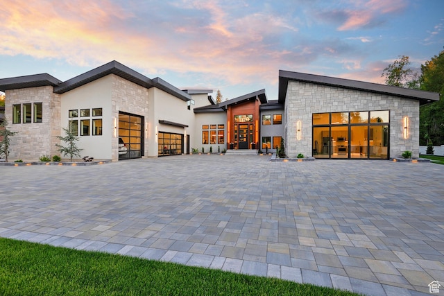 back of property at dusk with stone siding, stucco siding, an attached garage, and decorative driveway