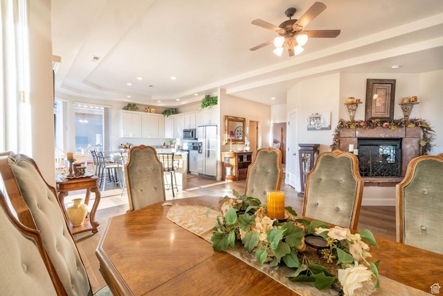 dining area with a tray ceiling, a multi sided fireplace, ceiling fan, and light wood-type flooring
