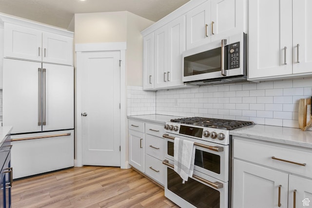 kitchen featuring light wood finished floors, high end appliances, light stone countertops, white cabinetry, and backsplash