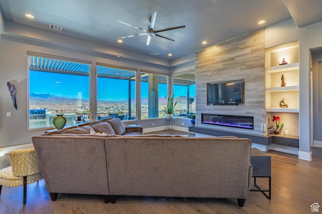 living room featuring a mountain view, built in features, ceiling fan, a tiled fireplace, and hardwood / wood-style floors
