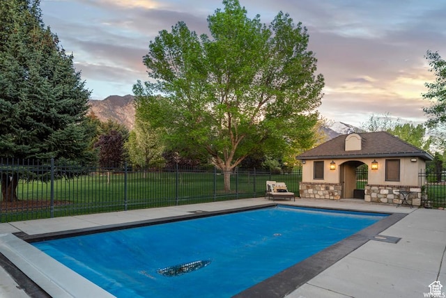 pool at dusk with a mountain view, a patio area, an outdoor structure, and a lawn