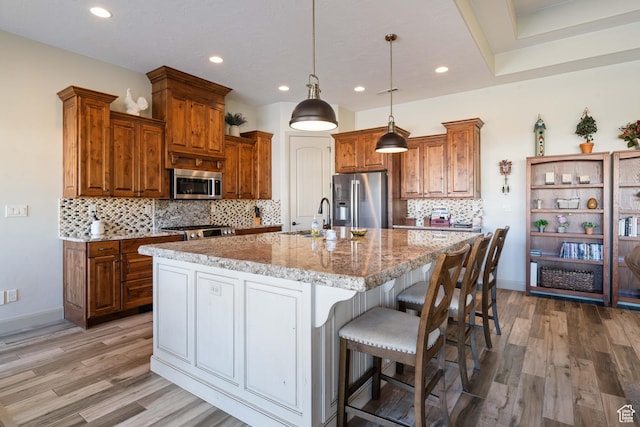 kitchen with sink, a kitchen island with sink, stainless steel appliances, light stone countertops, and decorative light fixtures