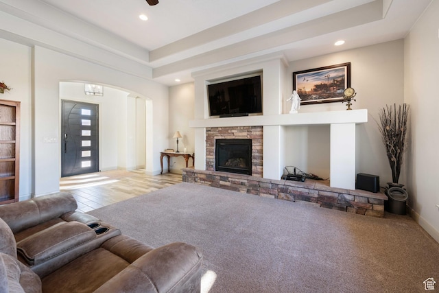 carpeted living room with a stone fireplace, a raised ceiling, and ceiling fan