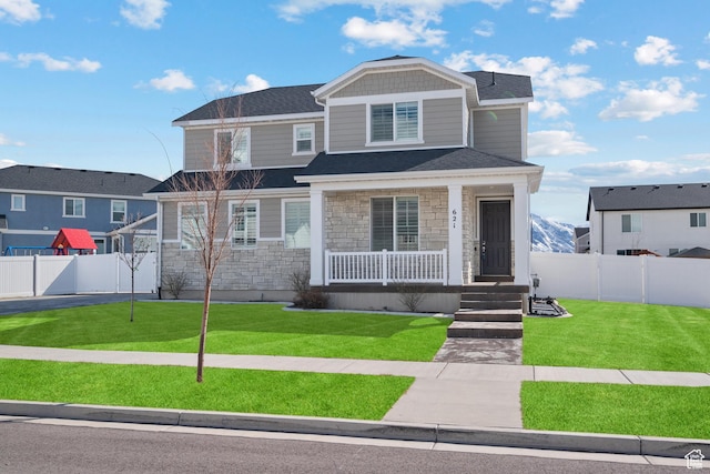 view of front facade featuring a front yard, roof with shingles, a fenced backyard, covered porch, and stone siding