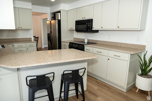 kitchen with light wood finished floors, a peninsula, light stone countertops, black appliances, and a kitchen bar