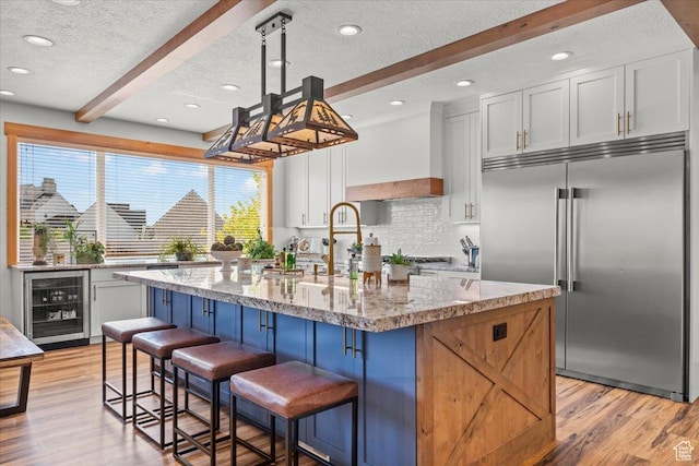 kitchen featuring beamed ceiling, light wood-style flooring, white cabinetry, wine cooler, and stainless steel built in fridge