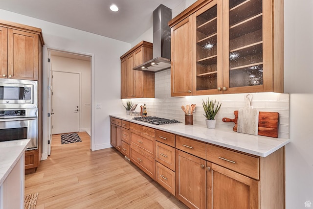 kitchen featuring wall chimney range hood, brown cabinetry, glass insert cabinets, and appliances with stainless steel finishes