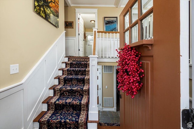 staircase featuring crown molding, a decorative wall, wainscoting, and visible vents
